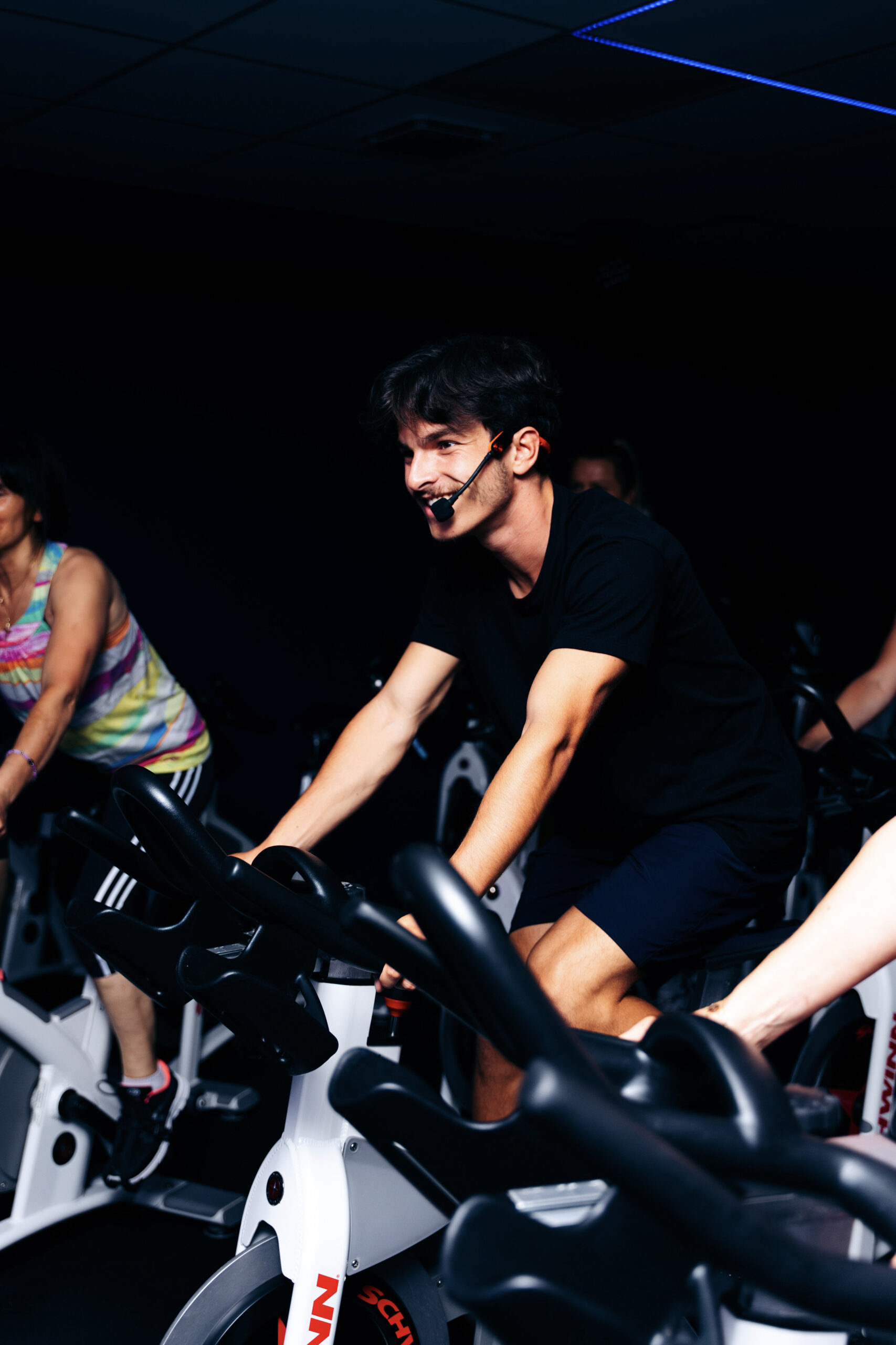 Instructeur souriant dirige un cours de vélo de spinning intense sur un Schwinn, portant un micro-casque, dans une salle sombre.