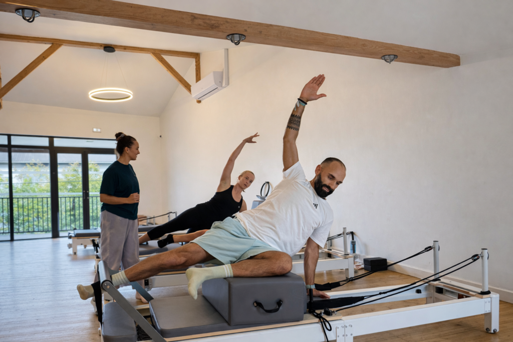 Entraînement Pilates : Deux élèves font une planche latérale sur reformers. Une instructrice les guide dans un studio lumineux.